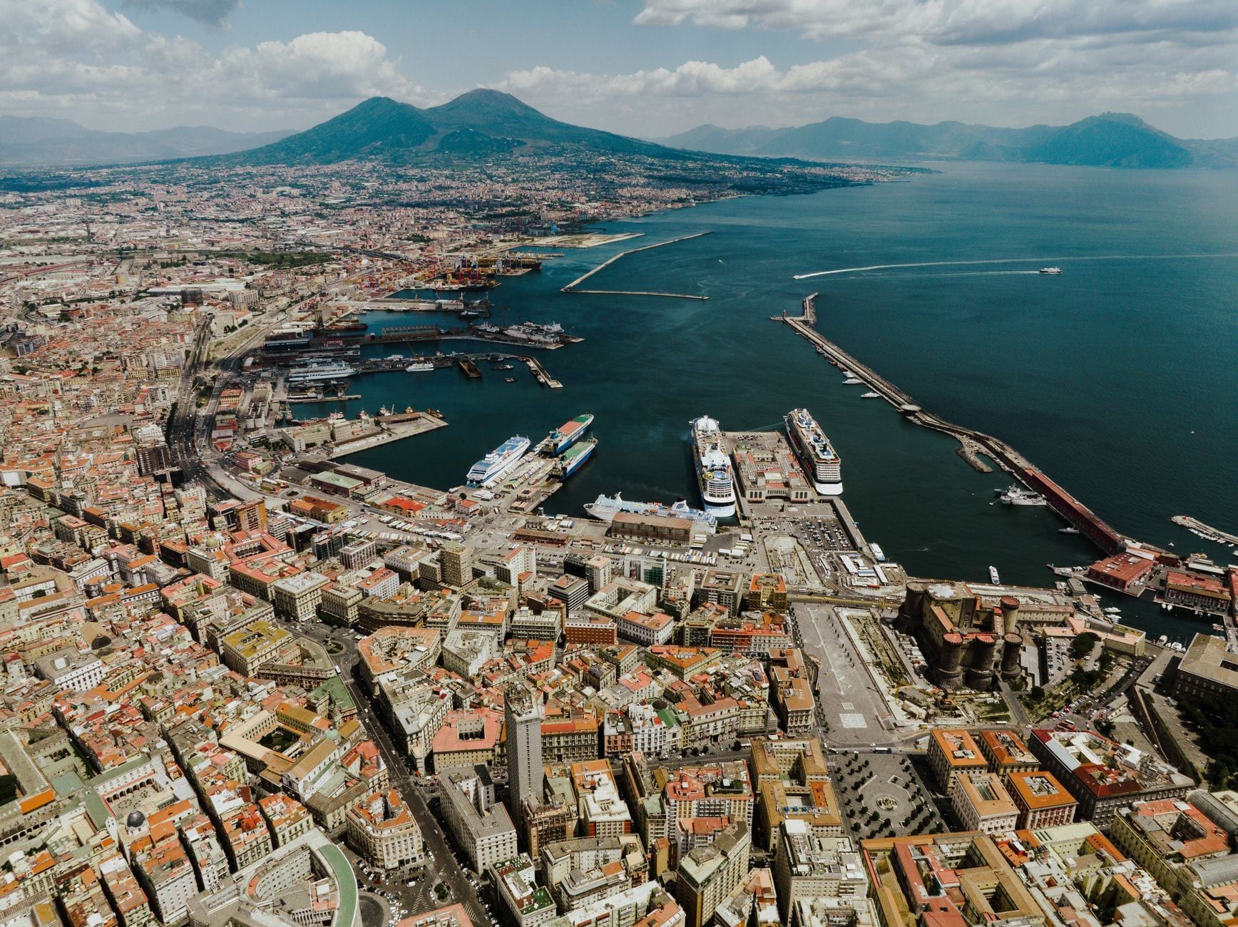 Aerial view over the city and harbor of Naples with Mount Vesuvius rising prominently in the background.