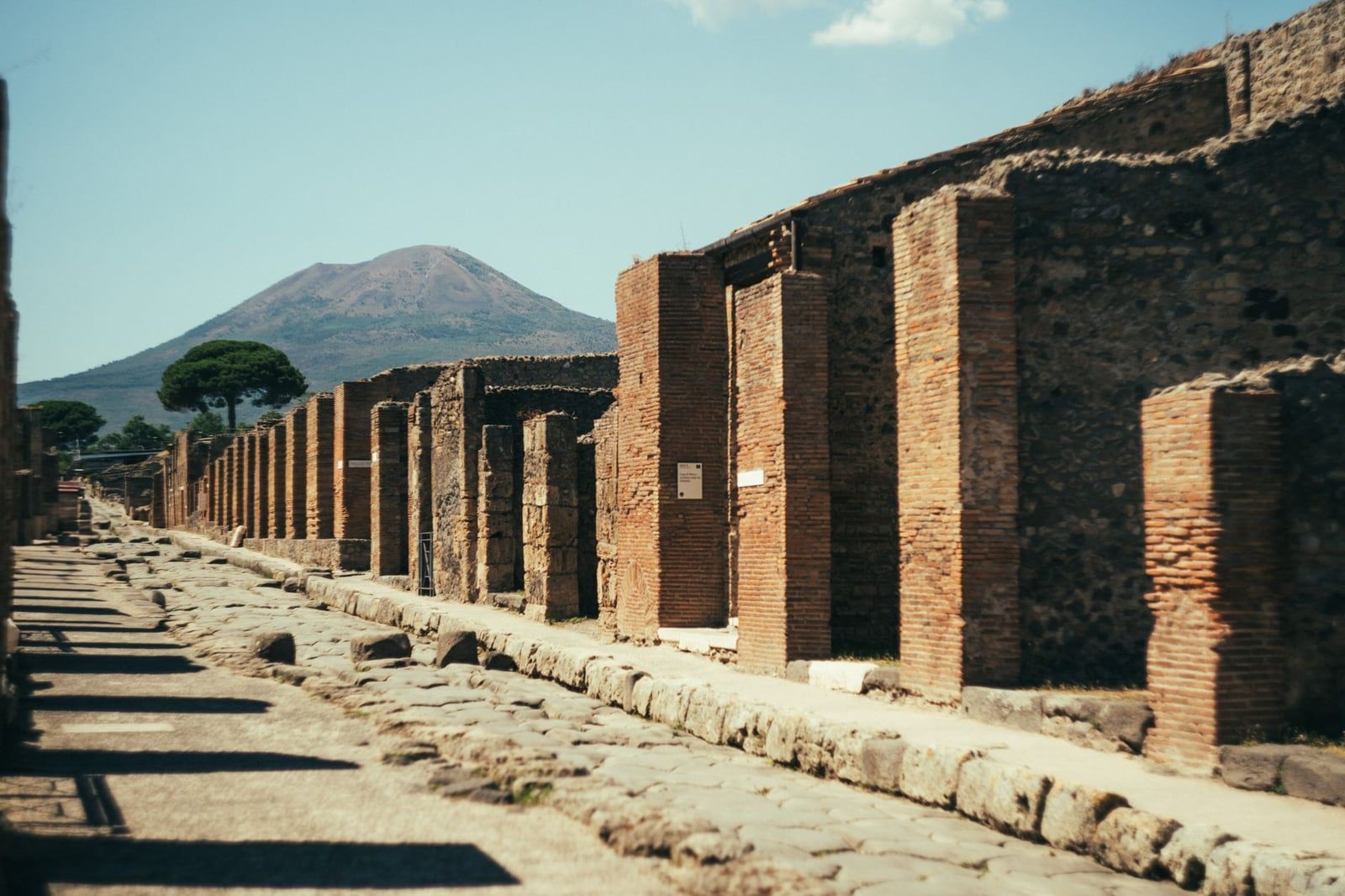 Ancient stone-paved street and ruined brick buildings in Pompeii with Mount Vesuvius rising in the background.